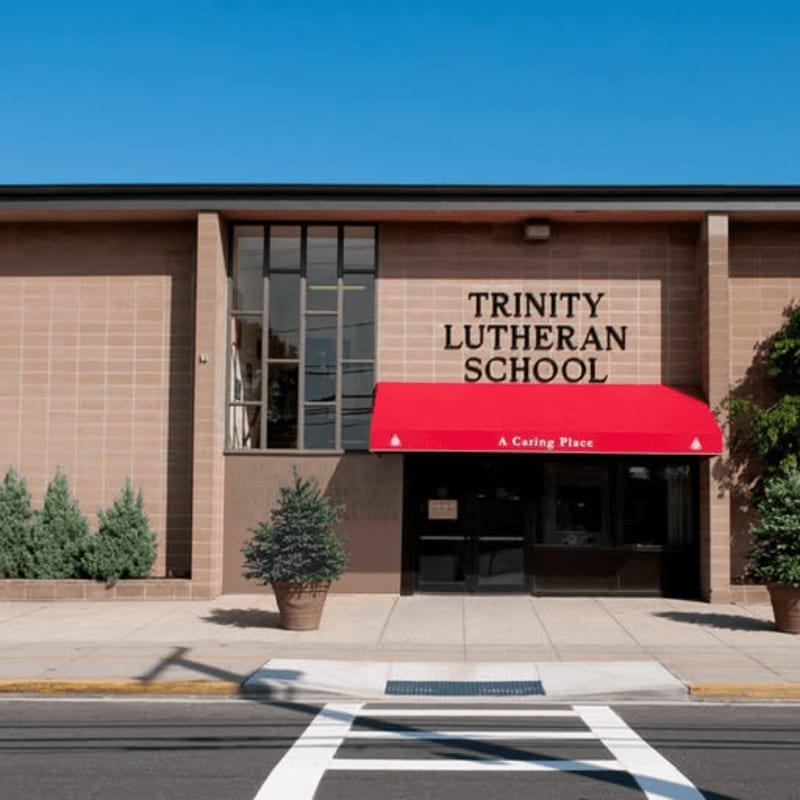 A red awning shades the entrance to Trinity Lutheran School, with a crosswalk in the foreground and a clear blue sky in the background.