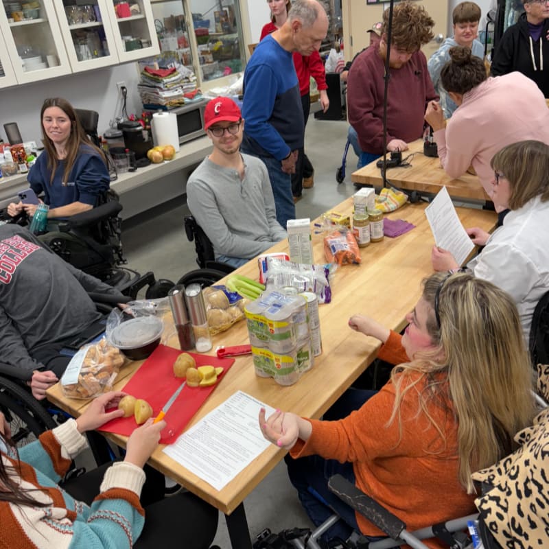 In the foreground, people in wheelchairs gather around a table with food and ingredients, while in the background, others stand and observe in a kitchen-like setting.