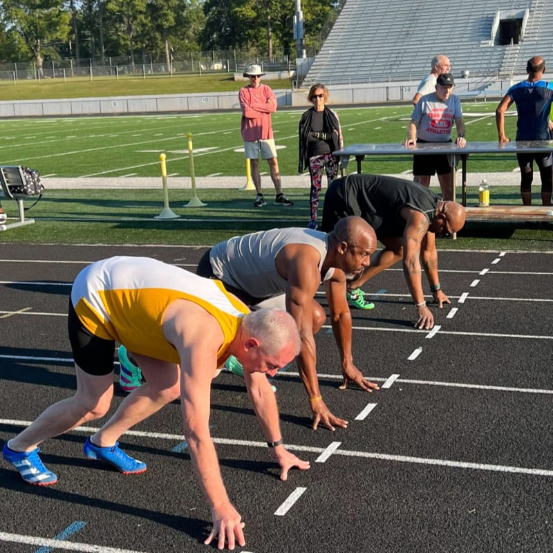 Athletes in starting positions on a track are in the foreground, while spectators watch from the bleachers in the background.
