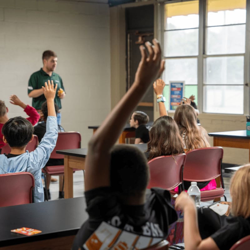 Students in a classroom raise their hands to answer a question from a teacher standing in front of them.