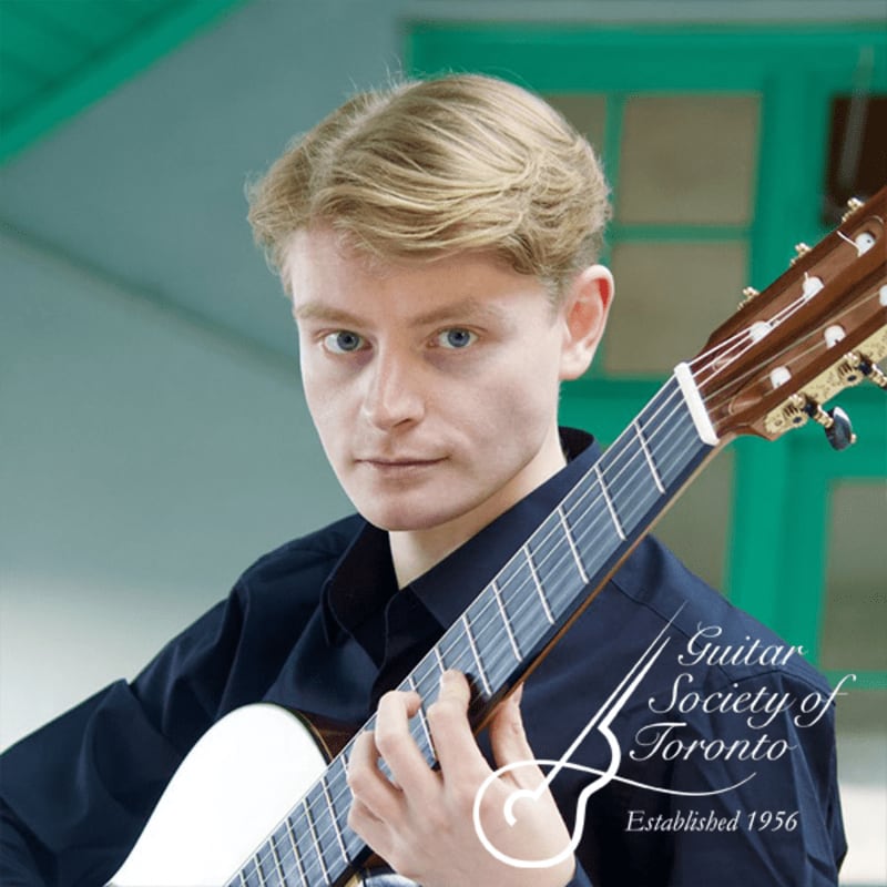 A young man with blond hair plays a guitar in front of a blurred background with a turquoise ceiling.