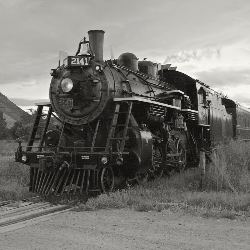 A vintage steam locomotive sits on tracks in tall grass, with a cloudy sky and distant hills in the background.