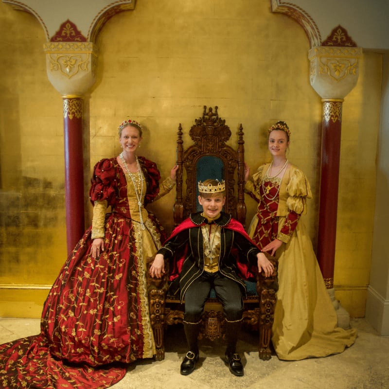 A young boy in royal attire sits on a throne between two girls in elaborate gowns, all against a shimmering gold backdrop with ornate pillars.