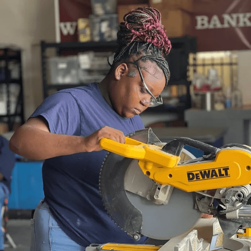 A young woman in a blue shirt and jeans, wearing safety glasses, operates a yellow miter saw in the foreground, while shelves with various items are visible in the blurred background.