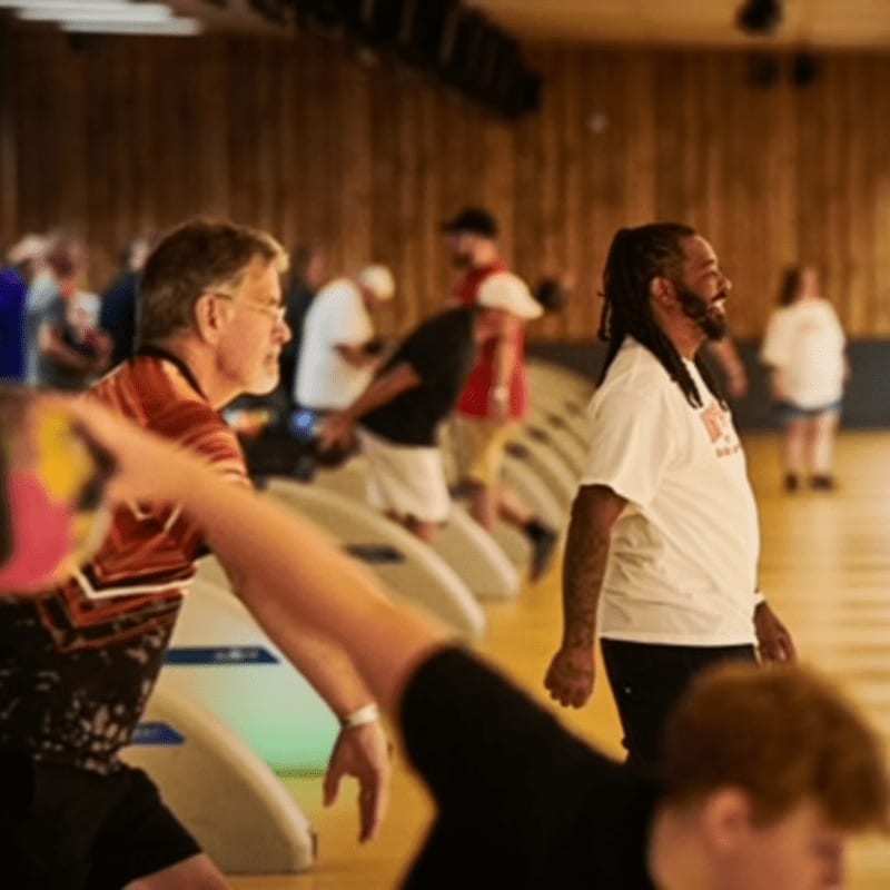 In the foreground, a man in a red shirt throws a bowling ball, while in the background, other bowlers and spectators are visible in a dimly lit bowling alley.