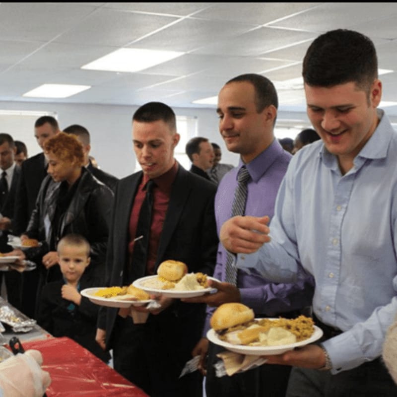 Men and a child in the foreground hold plates of food at a buffet line, while other people are visible in the blurred background.