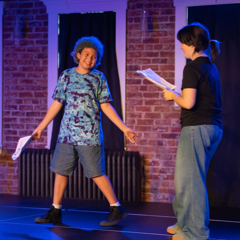 Two people stand on a stage with a brick wall and dark curtains in the background, while one person in a tie-dye shirt and shorts holds papers in the foreground.