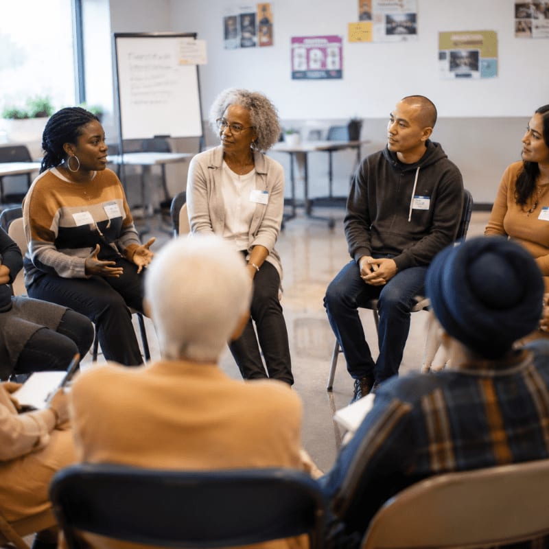 A diverse group of people sit in a circle in a brightly lit room with posters on the wall, engaged in conversation.