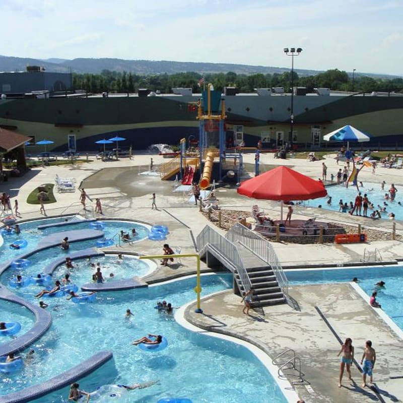 People enjoy a sunny day at a crowded water park with a lazy river in the foreground and a large building with a green and blue mural in the background.
