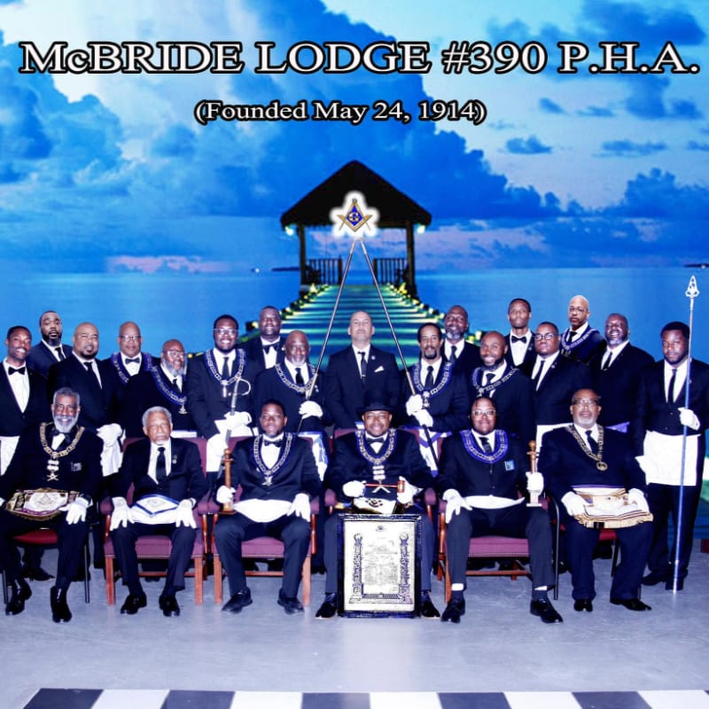 A group of men in formal attire are seated in the foreground, with a pier leading to a structure under a bright blue sky in the background.