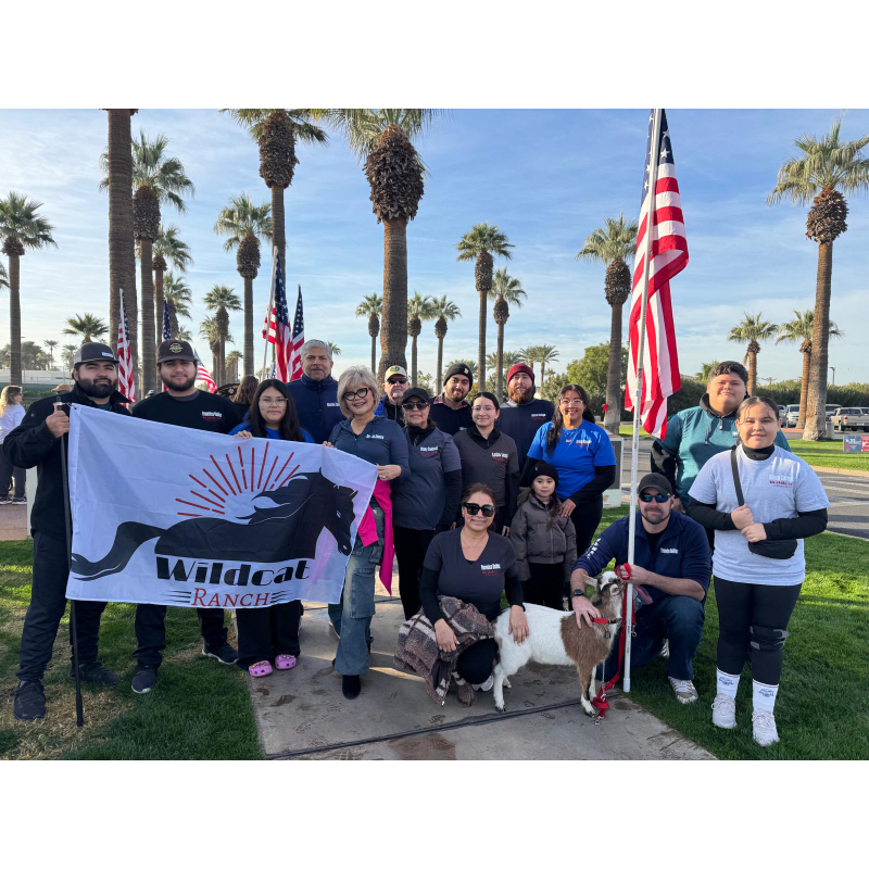 A group of people, some holding a flag and a goat, stand in front of palm trees and American flags.