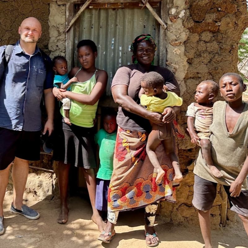A group of people, including a man in a blue shirt and shorts, stand in front of a mud-brick house with a corrugated metal door, while children of various ages are held or stand beside them.