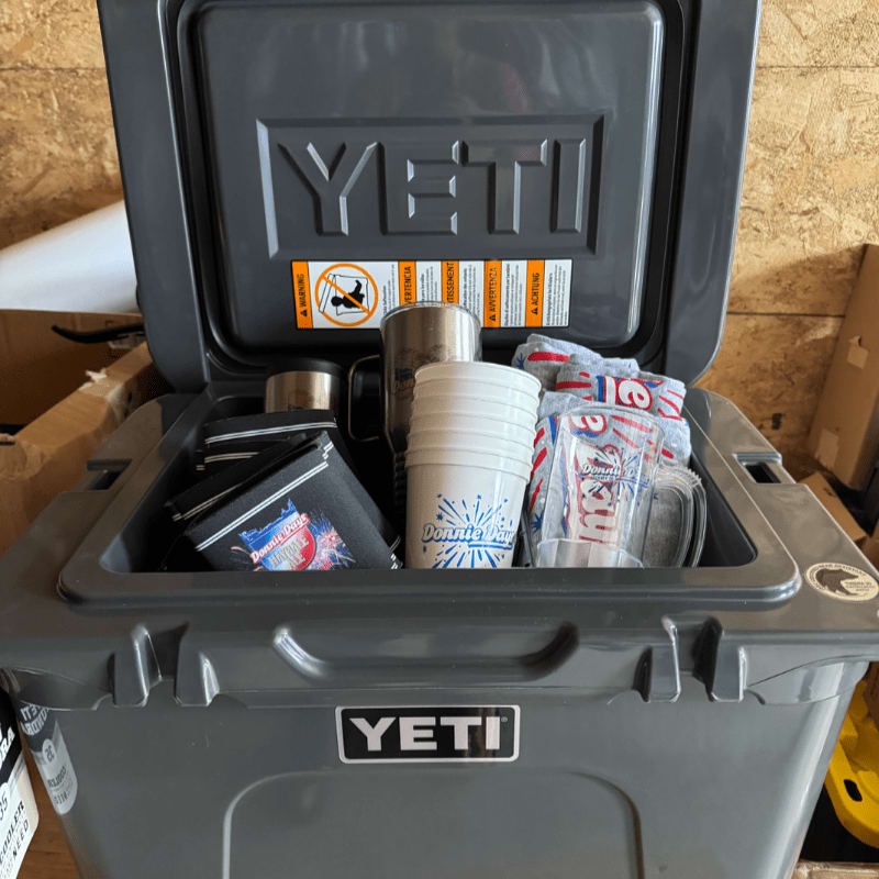 A gray YETI cooler is filled with cups and other items, with a wooden wall in the background.