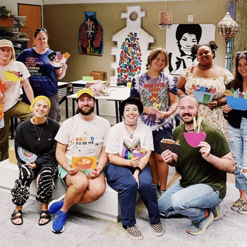 A group of smiling people, some holding colorful paper crafts, pose in a bright art studio with anatomical and artistic murals on the walls.