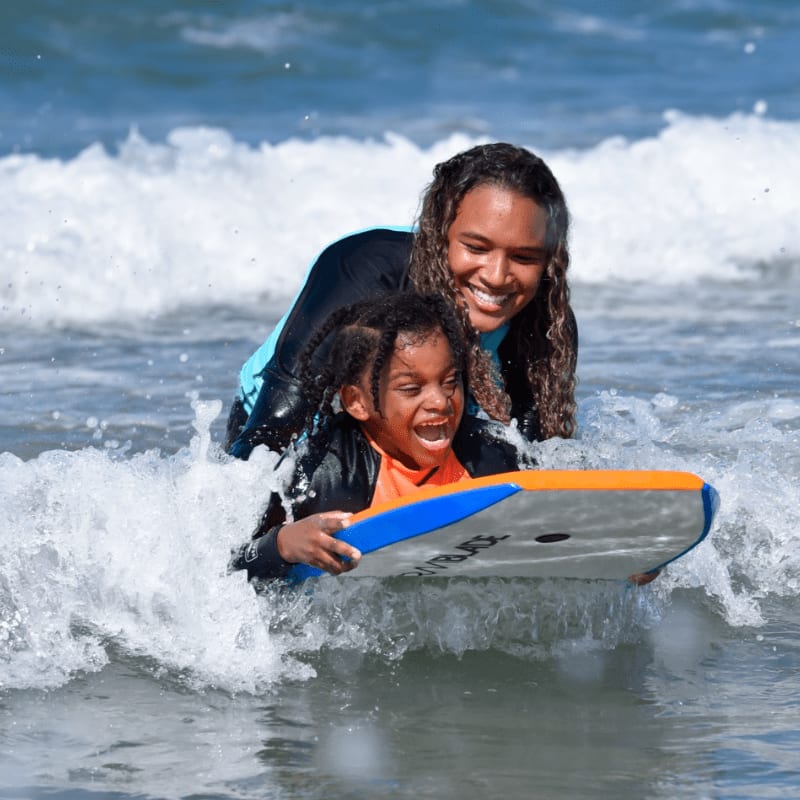 A woman and child ride a boogie board through a wave, with the ocean stretching out behind them.