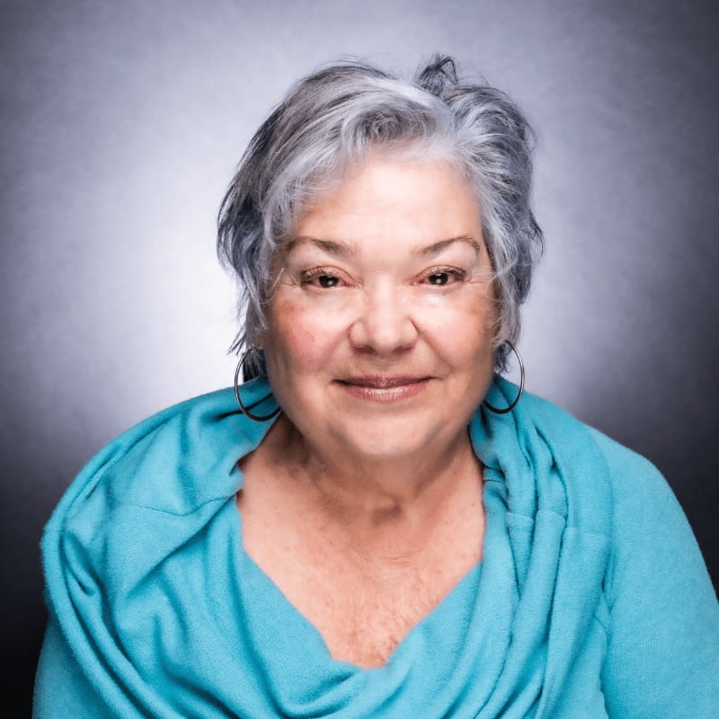 A smiling woman with gray hair and hoop earrings wears a teal shawl against a dark, gradient background.