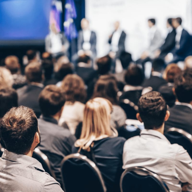 The foreground shows the backs of people's heads as they sit in chairs, while the background features a blurred panel of speakers on a stage.
