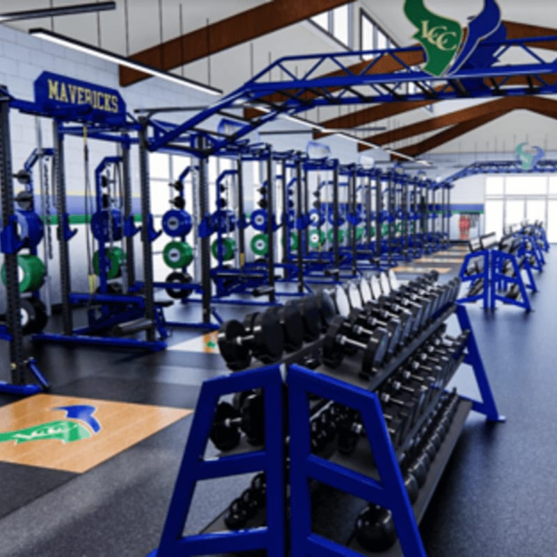 A row of dumbbells in the foreground leads to a spacious gym with weight racks and a "Mavericks" banner in the background.