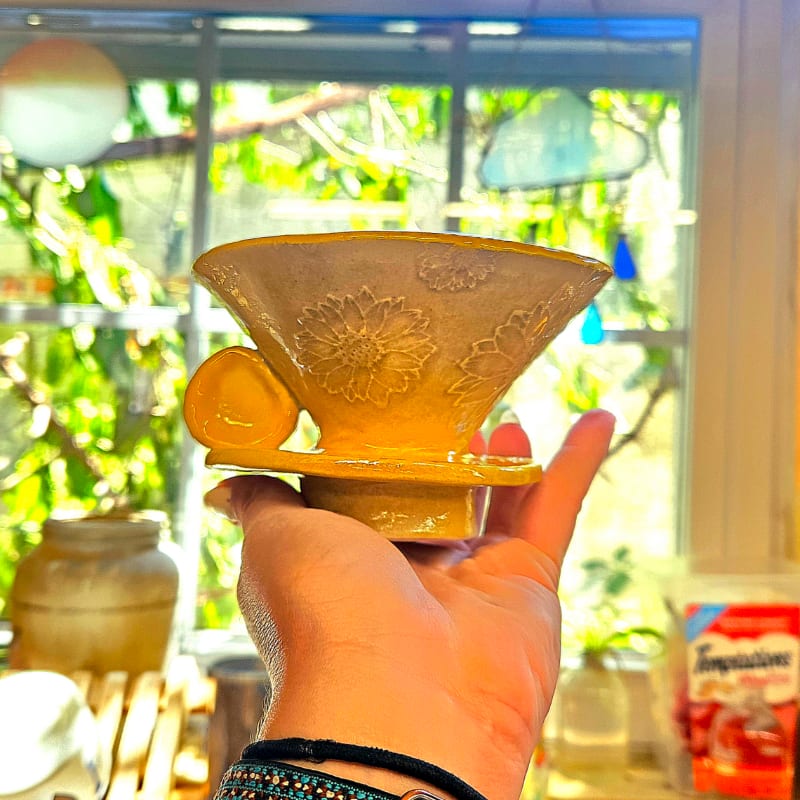 A hand holds a yellow ceramic coffee dripper with floral embossing in the foreground, while a sunlit window with greenery and kitchen items forms the background.
