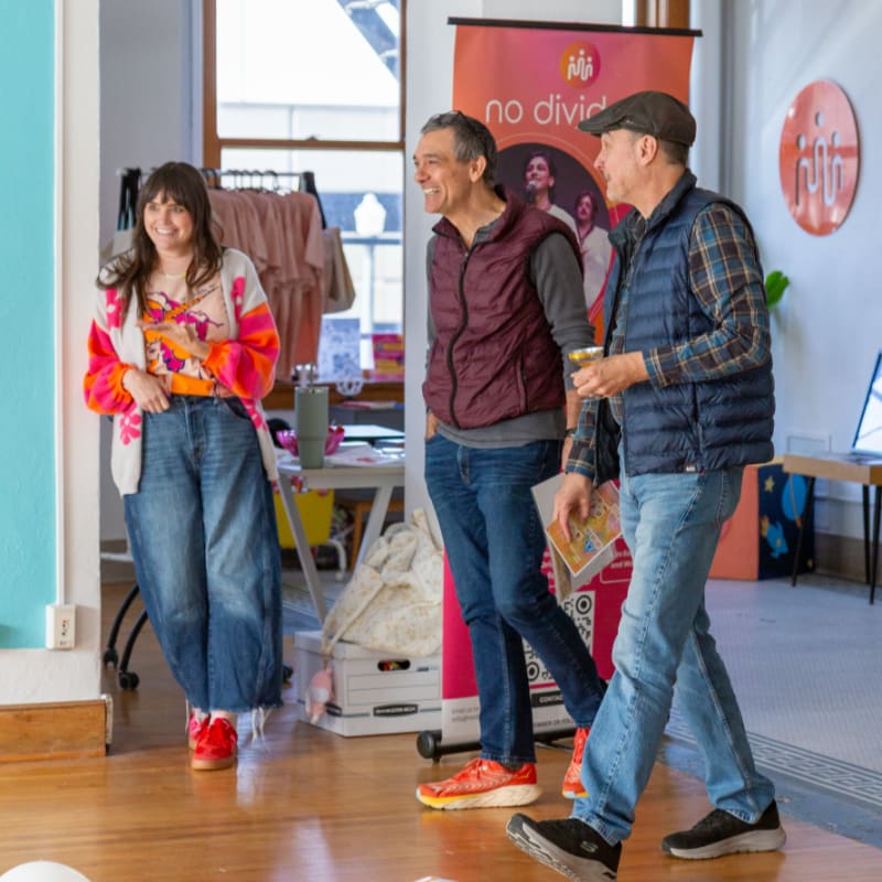 A woman in a colorful outfit stands to the left, while two men in casual attire walk past her, with a banner and clothing racks visible in the background.