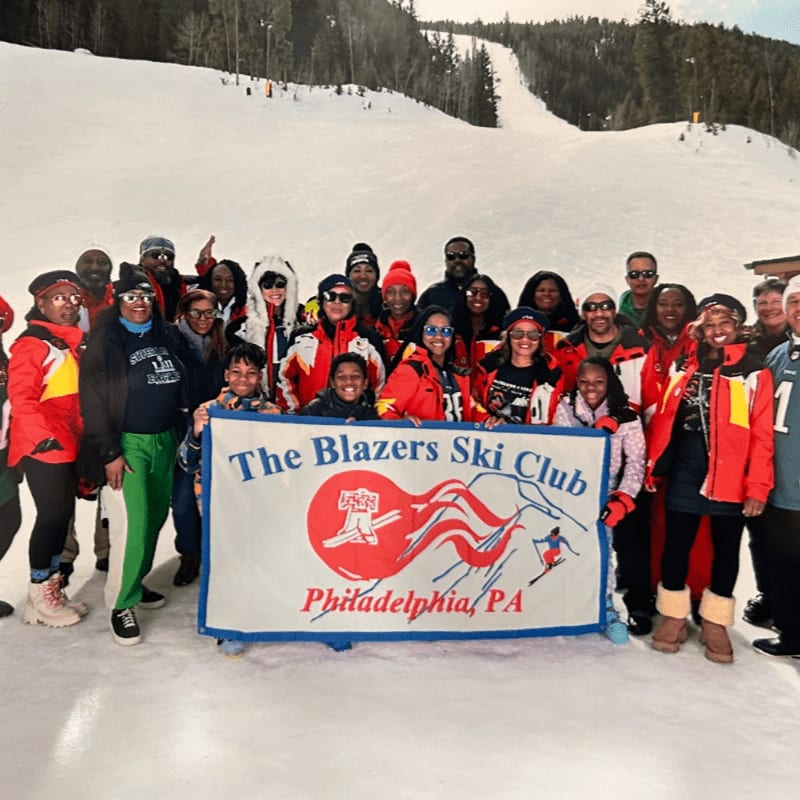 A large group of people in ski attire pose for a photo in front of a ski slope, holding a banner that reads "The Blazers Ski Club Philadelphia, PA."