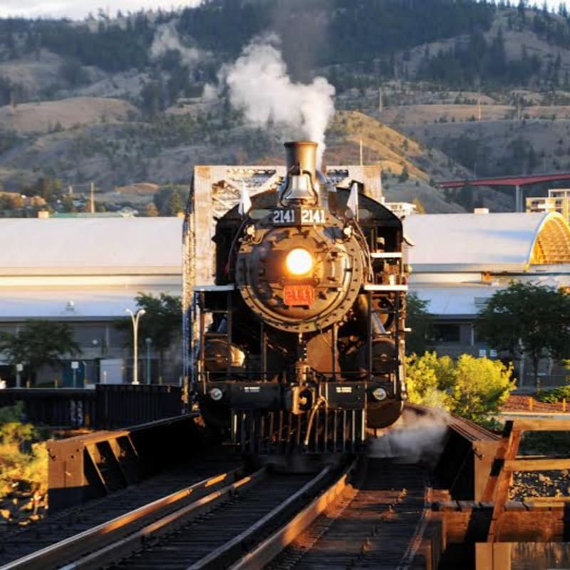 A vintage steam train with a glowing headlight chugs across a bridge with buildings and rolling hills in the background.