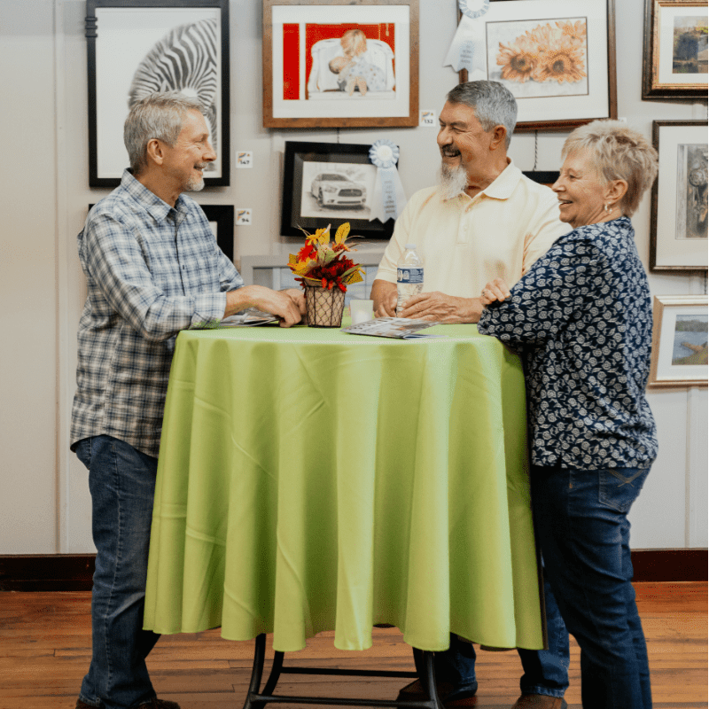 Three people stand around a lime green tablecloth-covered table in the foreground, while framed artwork adorns the walls in the background.