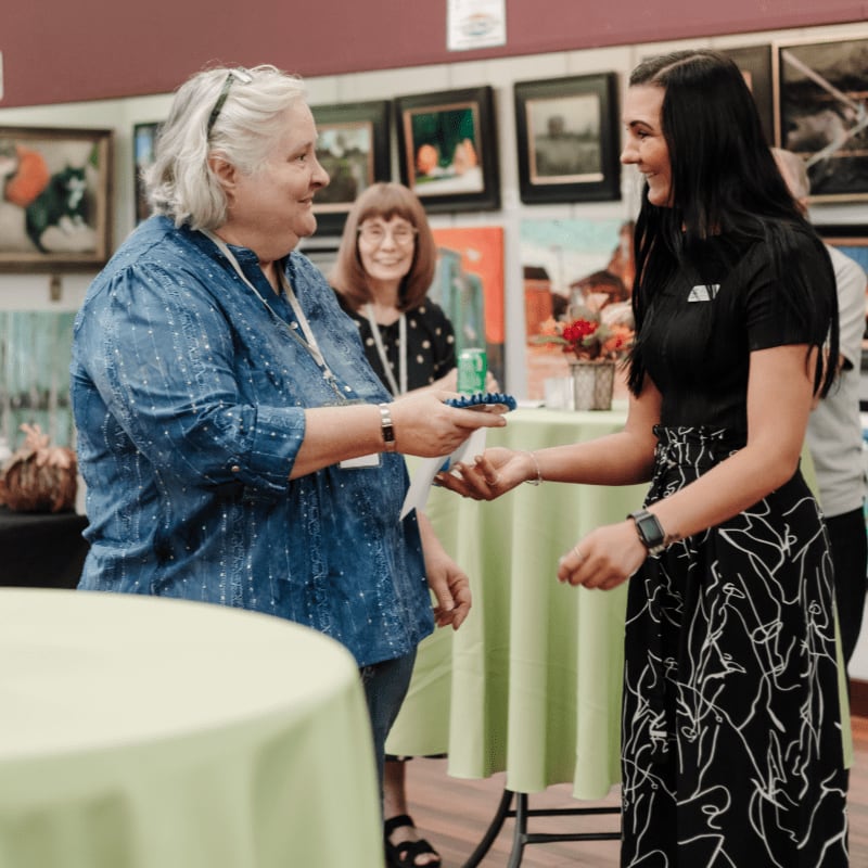 In the foreground, two women are interacting, with one handing a small item to the other, while the background features an art gallery with paintings on display.