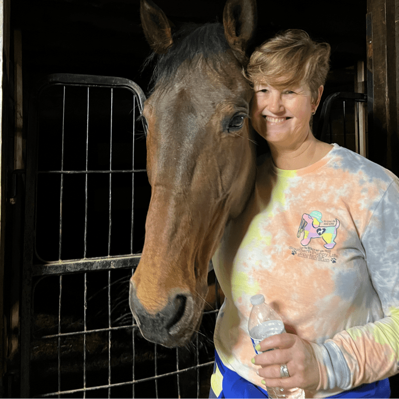 A woman with short blonde hair smiles as she poses with a brown horse in a stable, holding a water bottle.