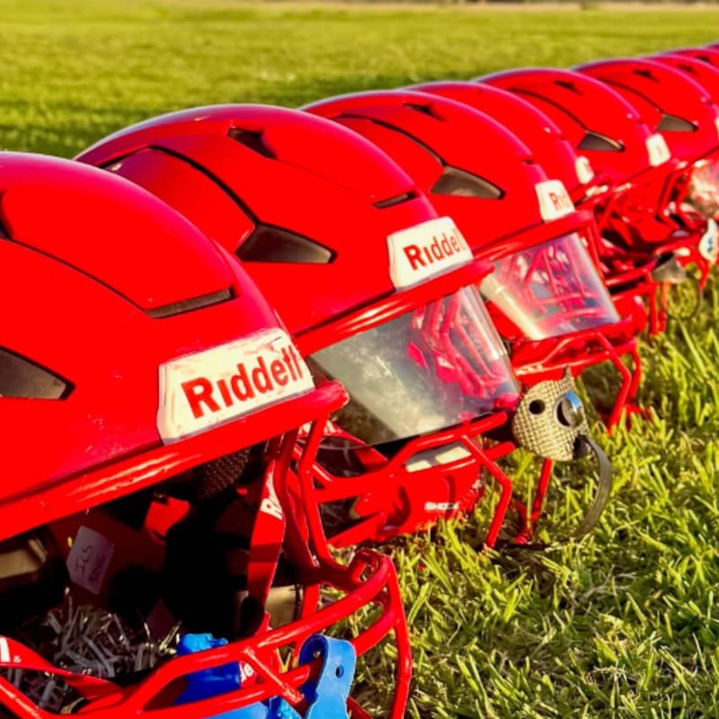 A row of red football helmets with visors sits on green grass, with a blurred grassy field and sky in the background.