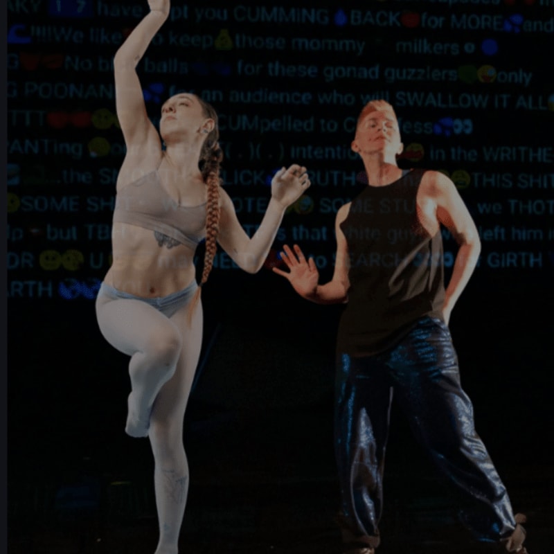 Two dancers, one in white leggings and a grey top, the other in a dark tank top and sparkly pants, perform on a dark stage with text projected behind them.