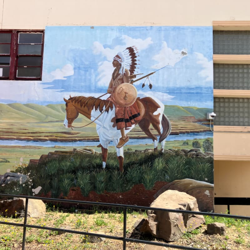 A Native American warrior on horseback stands in the foreground against a backdrop of a river valley and sky.