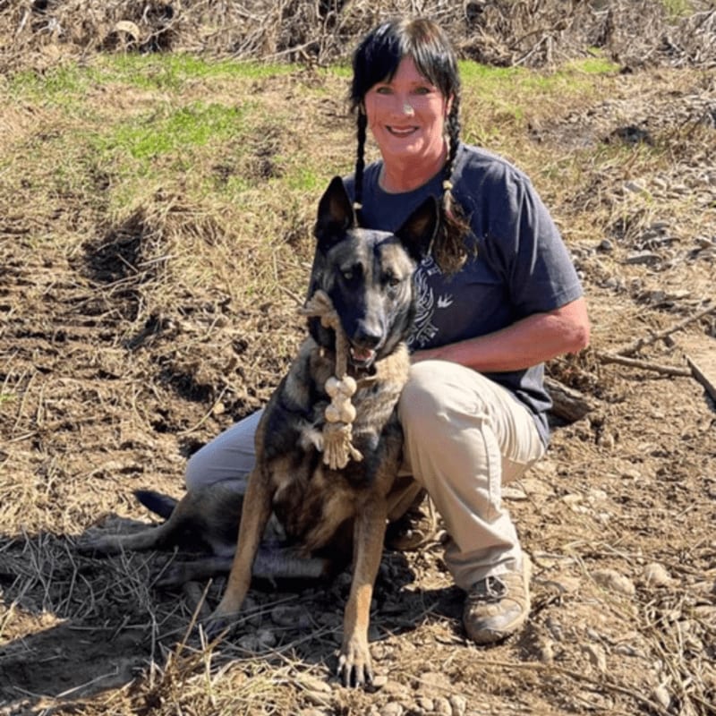 A woman with braided hair kneels beside her Belgian Malinois dog, which holds a rope toy, in a dry, grassy field.
