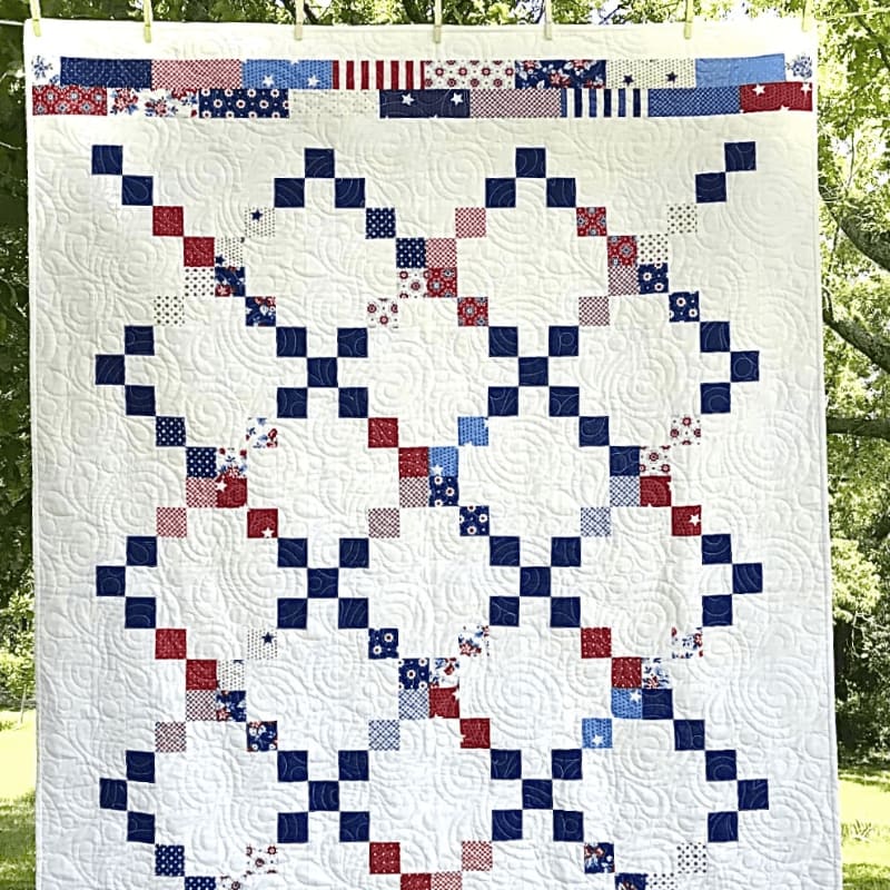A white quilt with a red, white, and blue checkered pattern hangs in front of a blurred green background of trees.