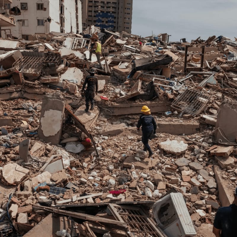 Rescuers in hard hats navigate a vast debris field of collapsed buildings under a hazy sky.