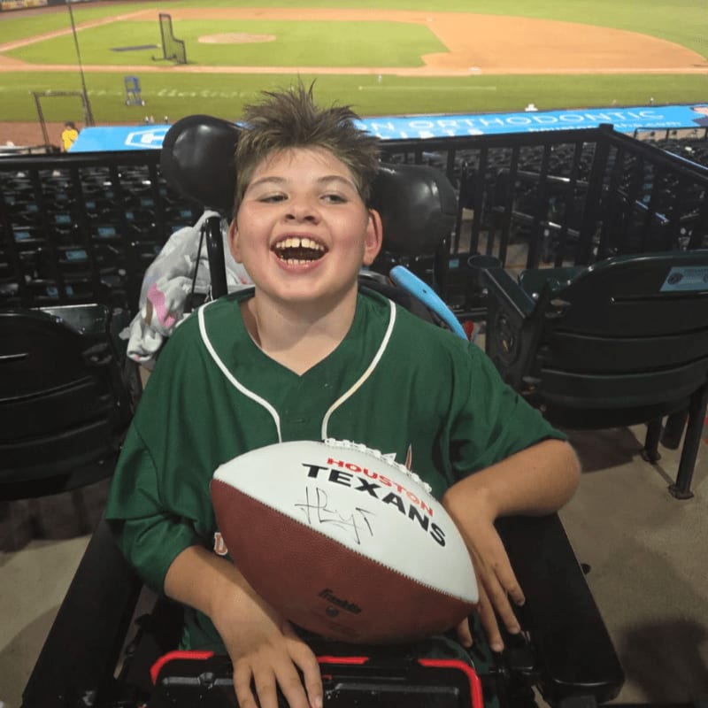 A smiling boy in a wheelchair holds a football in front of a baseball stadium.