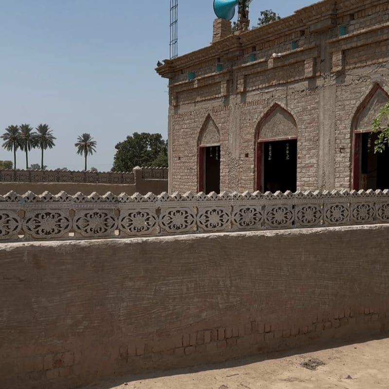A textured foreground wall leads to a decorative stone railing in front of a brick building with arched doorways, set against a backdrop of palm trees and a clear blue sky.