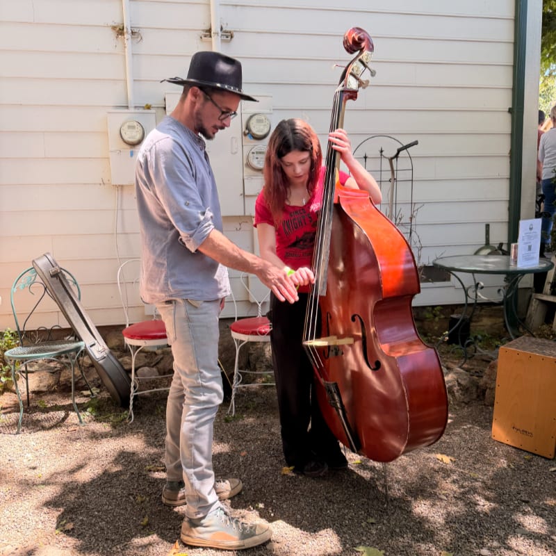 A man in a hat and a young woman stand by a double bass in a sun-dappled outdoor area with a white building and tables in the background.