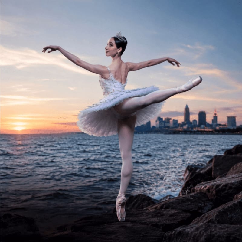A ballerina in a white tutu poses on rocky shore with a city skyline visible across the water at sunset.