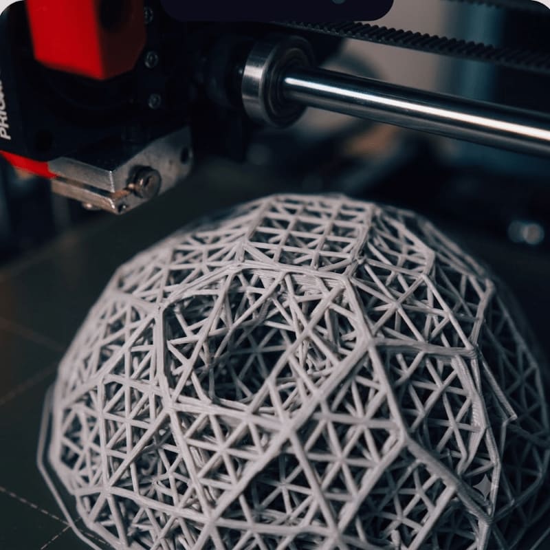 A 3D printer nozzle hovers over a complex, geodesic dome structure being printed in the foreground, with the printer's mechanical components visible in the background.