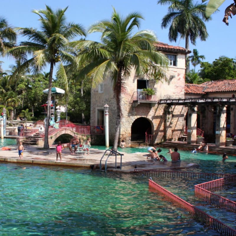 People swim and relax in a vibrant turquoise pool in the foreground, while a historic stone building with a tower and lush palm trees form the background.
