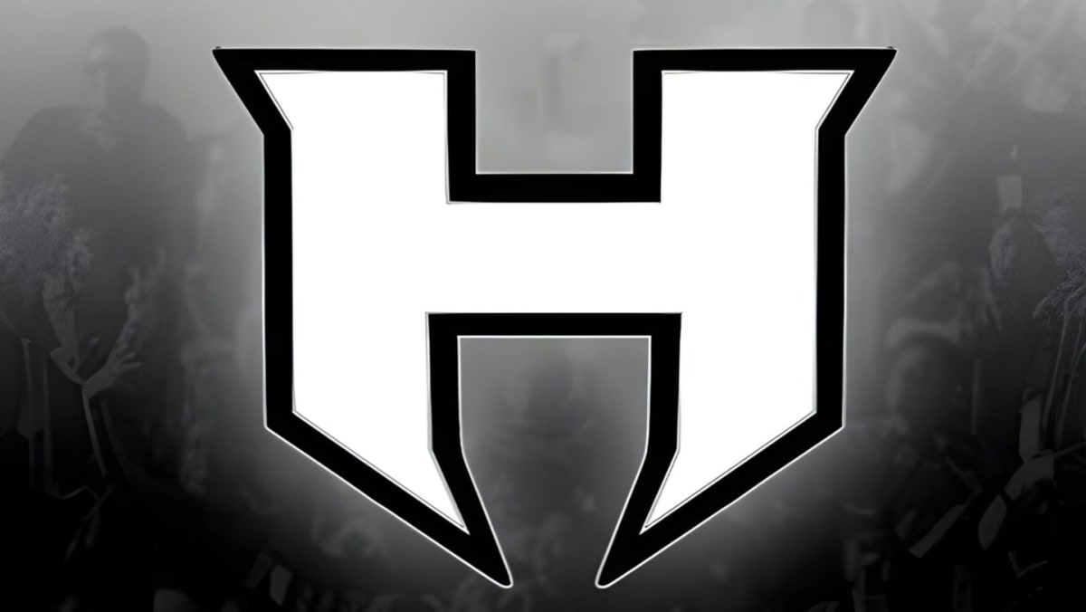 A large white letter "H" with a black outline dominates the foreground, set against a blurred, shadowy background of what appears to be a crowd of people.