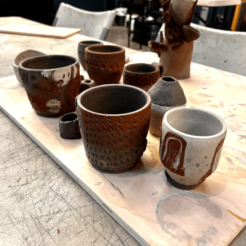 Several handmade ceramic cups and vases with textured surfaces are arranged on a wooden table, with a blurred background of a studio space.