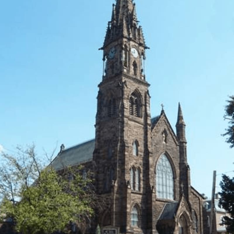 A tall, stone church with a clock tower stands against a clear blue sky, with green trees in the foreground.