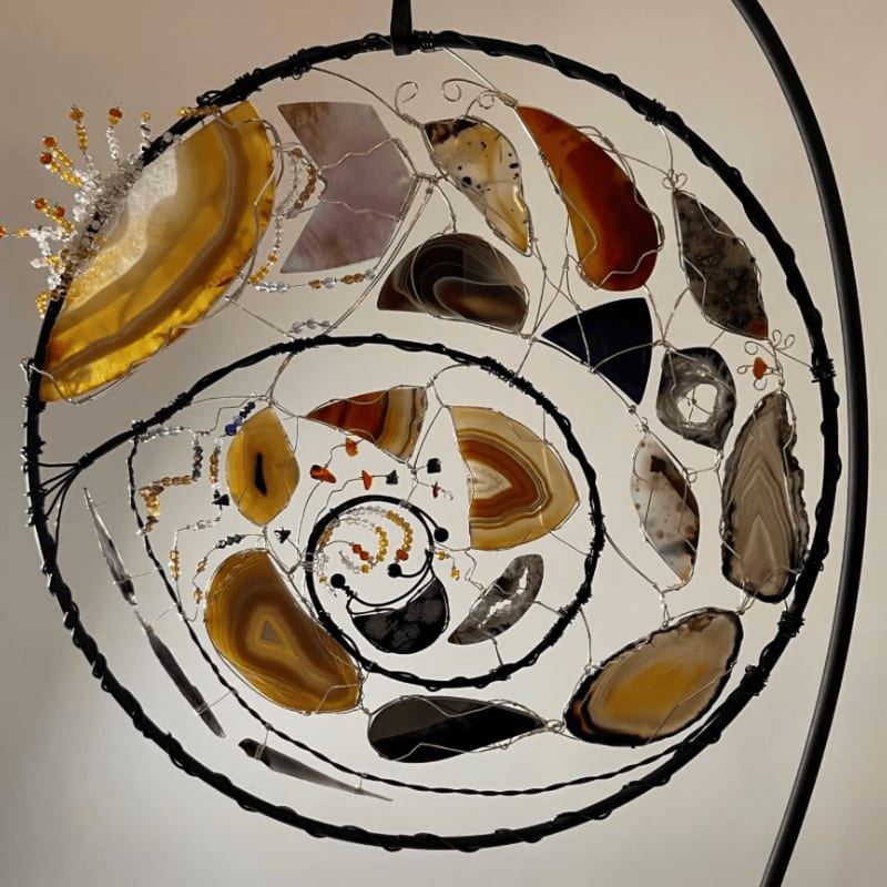 A circular, wire-wrapped dreamcatcher adorned with sliced agate stones and beads hangs against a plain, light-colored background.