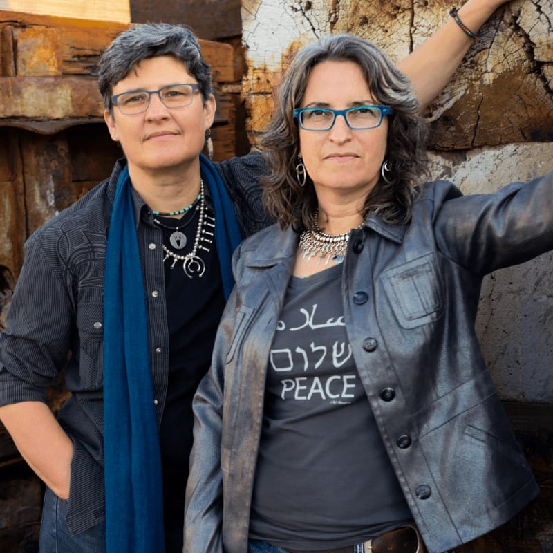 Two women stand in front of a rustic, textured wall, one wearing a blue scarf and the other a leather jacket over a shirt with "shalom peace" written on it.