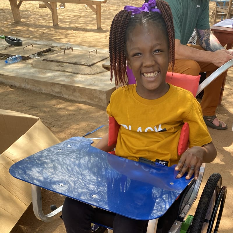 A smiling young girl in a wheelchair with a blue tray in front of her sits outdoors with a person in the background.