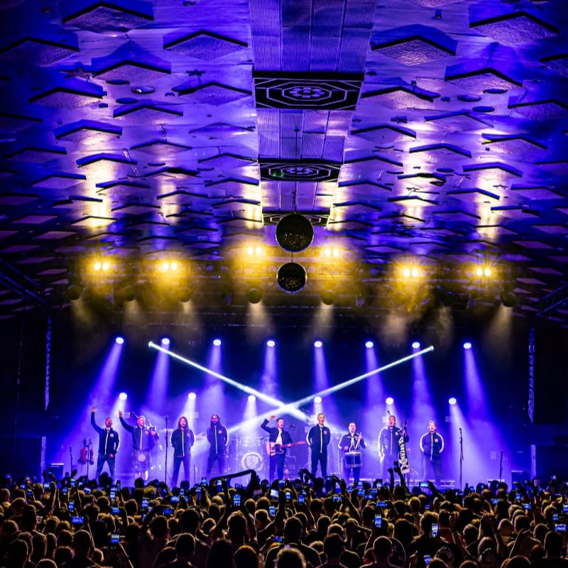 A band performs on a brightly lit stage in front of a large, cheering crowd, with a uniquely patterned ceiling illuminated by purple lights above.