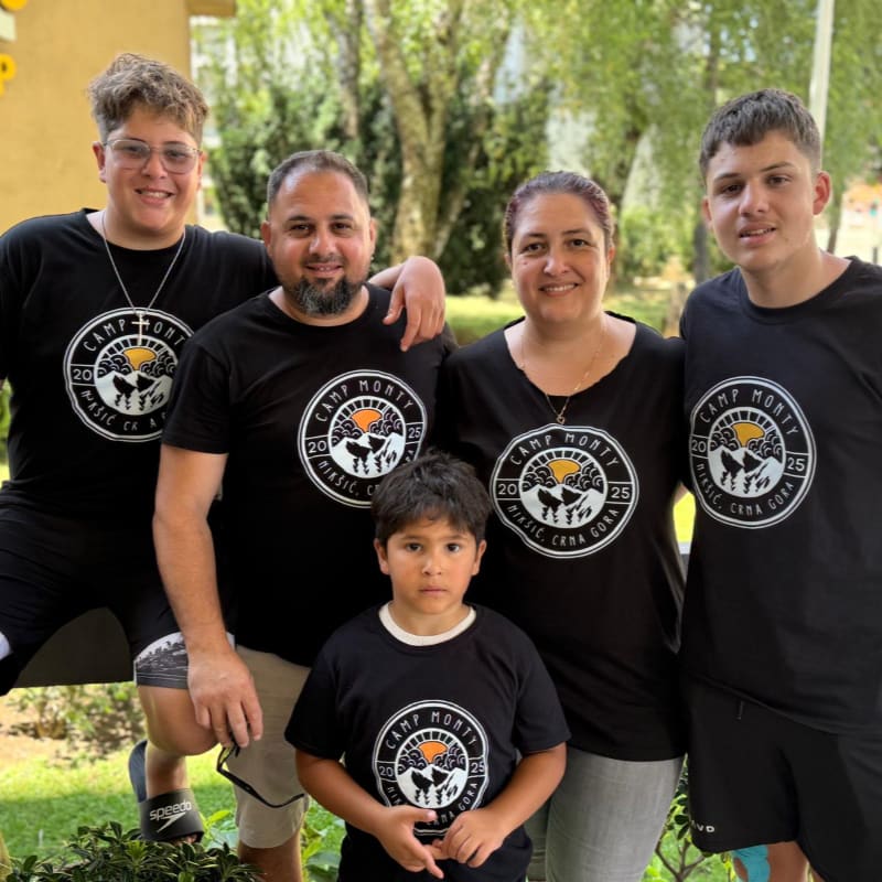 A family of five, all wearing matching black t-shirts with a circular logo, poses for a photo outdoors with lush green foliage in the background.