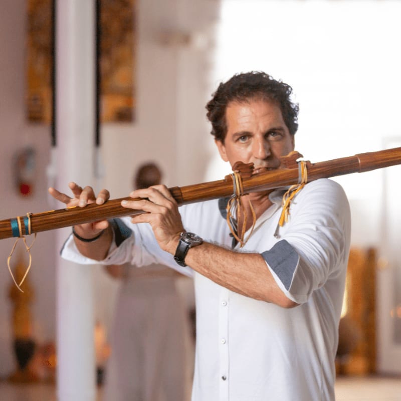 A man in a white shirt plays a wooden flute in the foreground, while a blurred figure stands in the background of a bright, airy room.